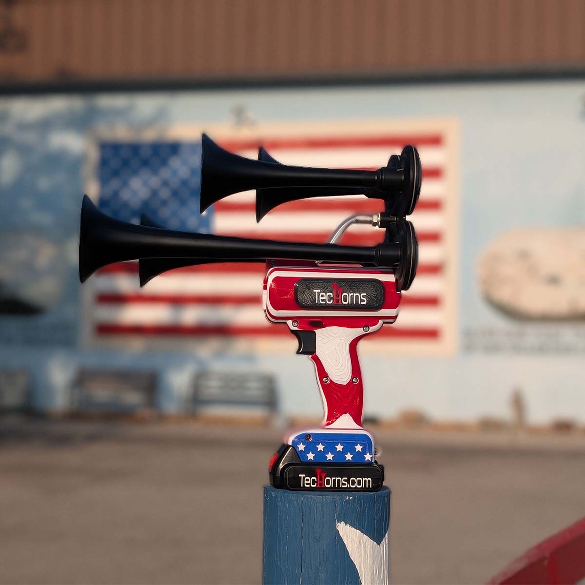 TecHorns ‘Merica 4-Horn – handheld train horn with 20 V lithium battery, displayed in front of a large American flag mural highlighting its red, white, and blue patriotic design