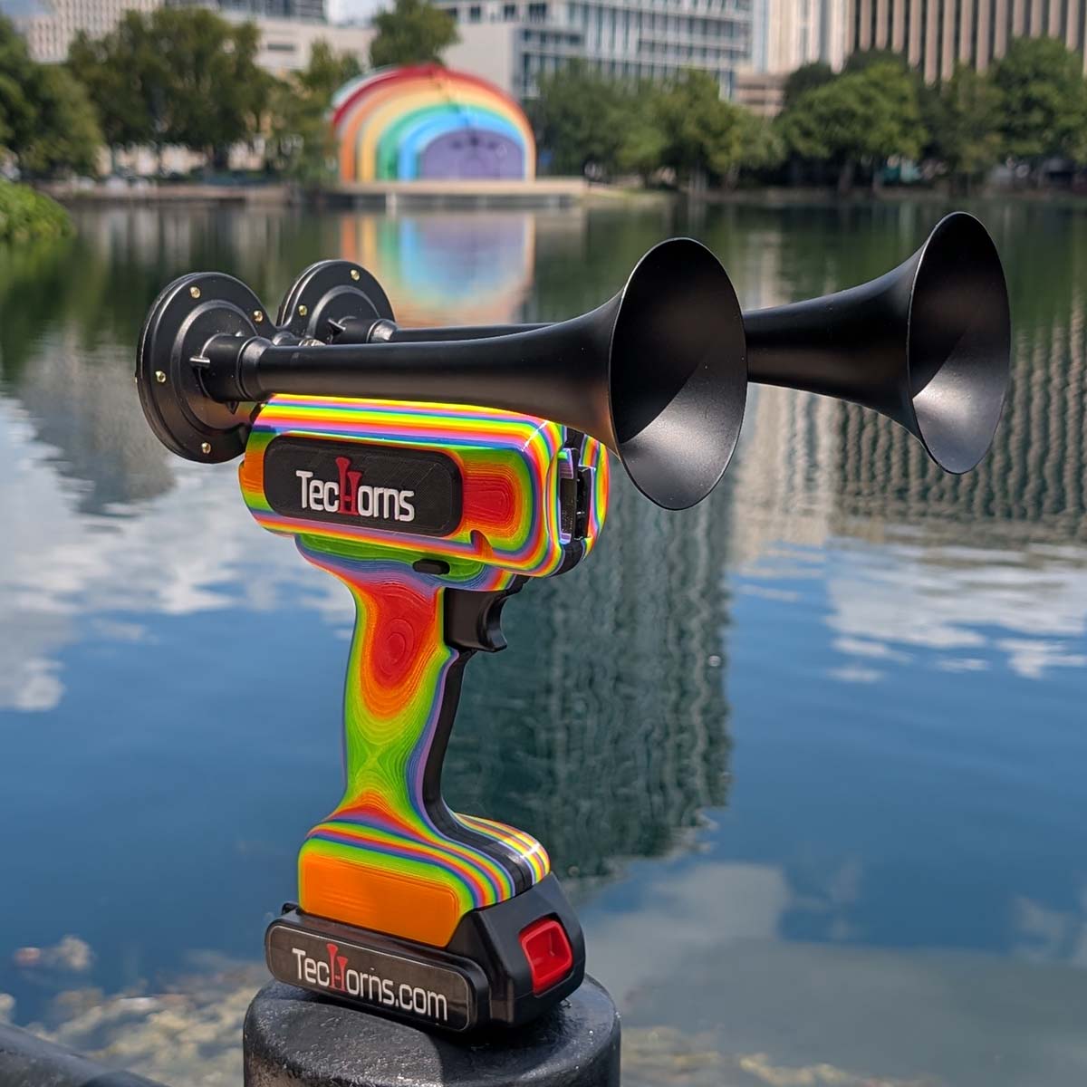 TecHorns RAINBOW 2-Horn – handheld train horn with 20 V lithium battery, featuring black trumpets and a rainbow-fade body, displayed in front of the Rainbow Amphitheater overlooking Lake Eola in Orlando, Florida