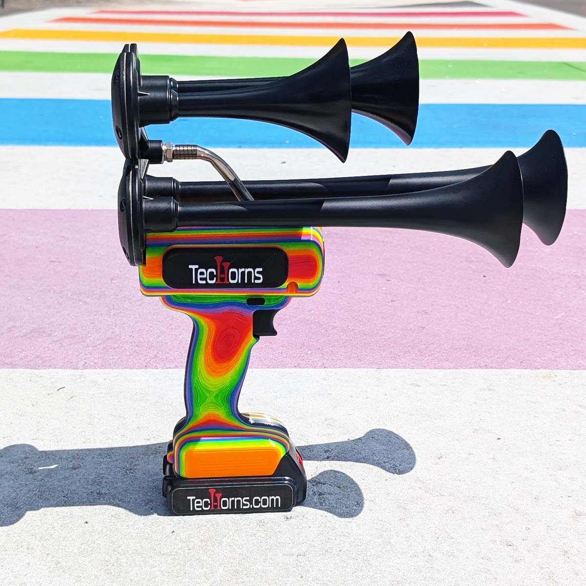 TecHorns RAINBOW 4-Horn – handheld train horn with 20 V lithium battery, featuring black trumpets and a bright rainbow-fade body, displayed in front of a colorful rainbow sidewalk