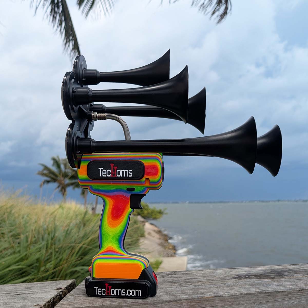 TecHorns RAINBOW 5-Horn – handheld train horn with 20 V lithium battery, featuring black trumpets and a vibrant rainbow-fade body, photographed outdoors near a river with storm clouds and palm trees in the background