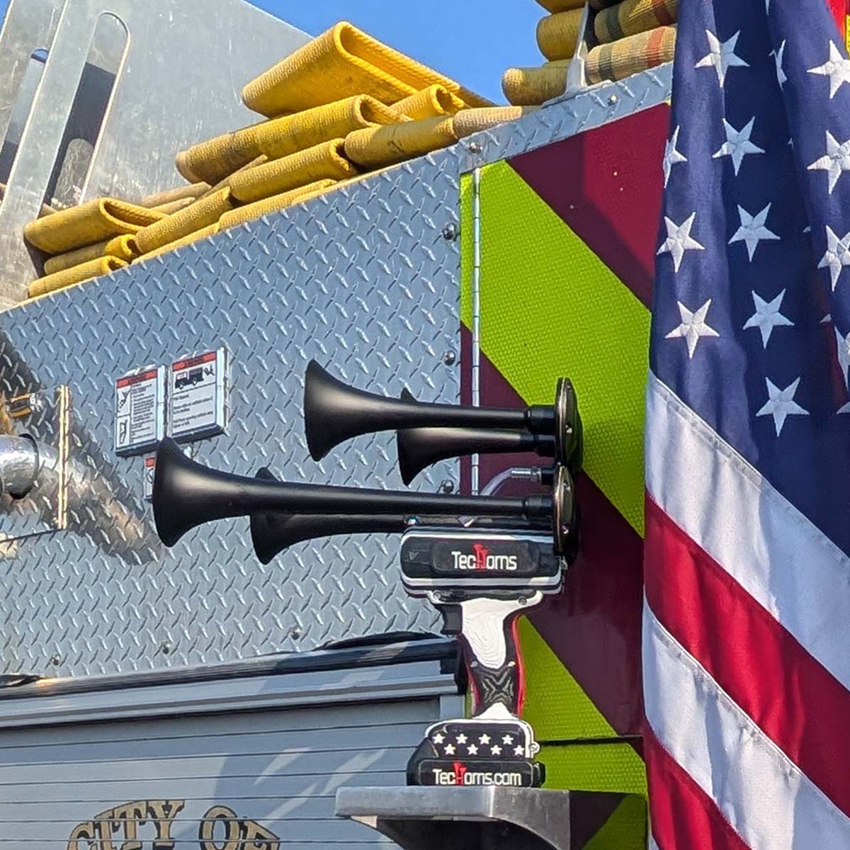 Thin RED Line TecHorns 4-Horn – handheld train horn with 20 V lithium battery, displayed on the chrome bumper of a red fire truck, reflecting its black-and-white design with the Thin RED Line stripe that honors firefighter courage and sacrifice.