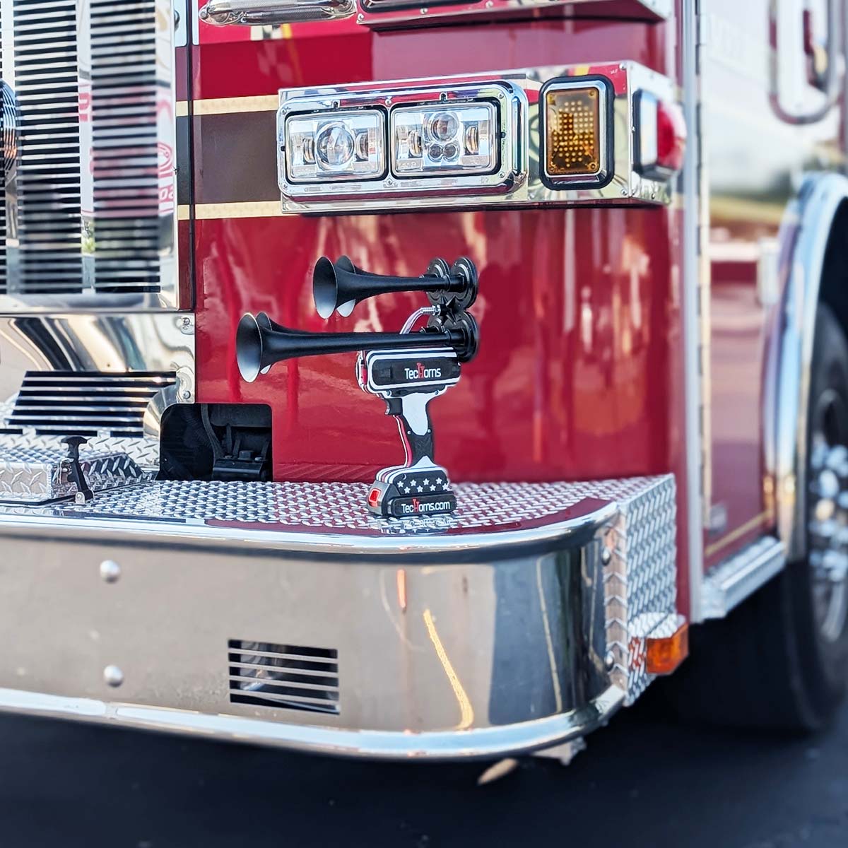 Thin RED Line TecHorns 4-Horn – handheld train horn with 20 V lithium battery, displayed on the chrome front bumper of a fire truck with red paint and gold striping, honoring firefighters and the Thin RED Line of courage
