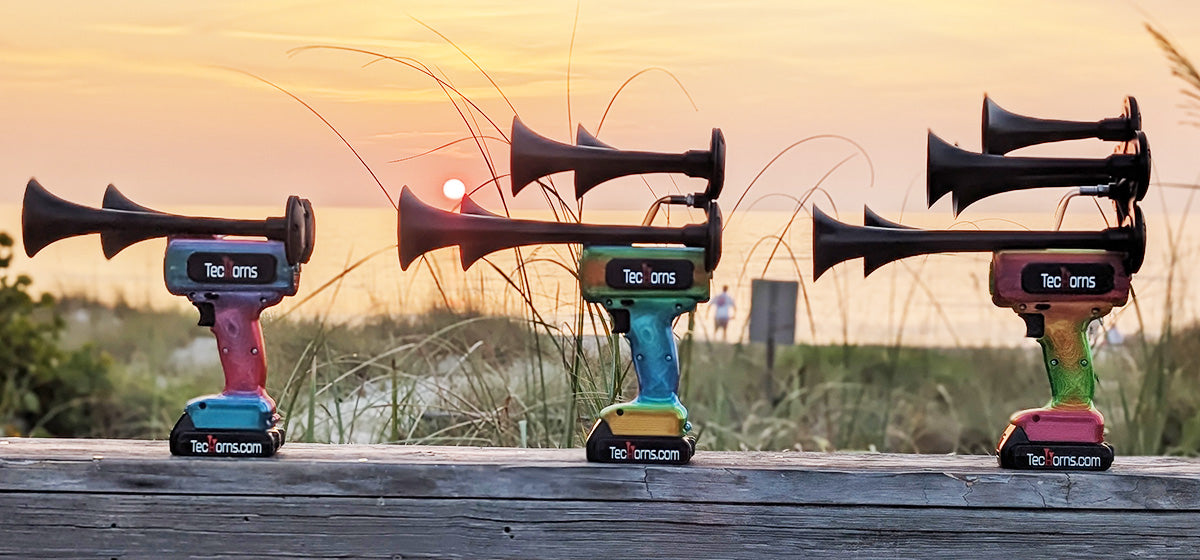TecHorn Candy Series lineup – Fire & Ice 2-Horn, Mango Bay 4-Horn, and Tropical Blaze 5-Horn handheld train horns with 20 V lithium batteries, displayed on a wooden rail at the beach during sunset
