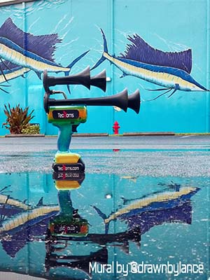 Mango Bay TecHorn 4-Horn handheld train horn with 20 V lithium battery reflected in a puddle beneath a vibrant sea life mural by @drawnbylance, showcasing custom color design and creative photography