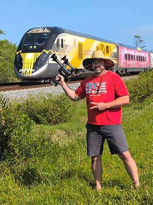 Man wearing a USA shirt holding a 'Merica TecHorn 4-Horn handheld train horn with 20 V lithium battery near a passing high-speed train, representing American pride and TecHorn power