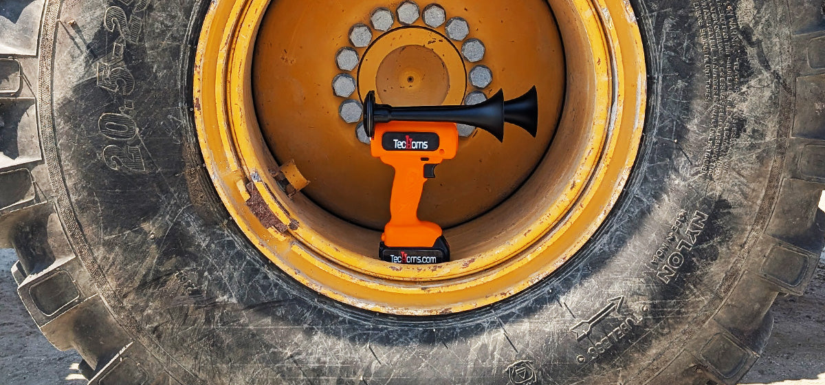 TecHorn Orange 2-Horn – handheld train horn with 20 V lithium battery and black trumpets, displayed inside a large yellow heavy equipment tire, highlighting its rugged power and industrial strength