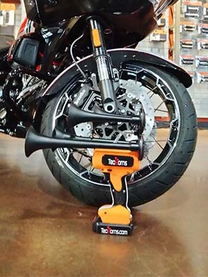 Orange, white, and black TecHorn 4-Horn handheld train horn with 20 V lithium battery displayed in front of a custom motorcycle front wheel, showcasing bold color design and high-performance craftsmanship