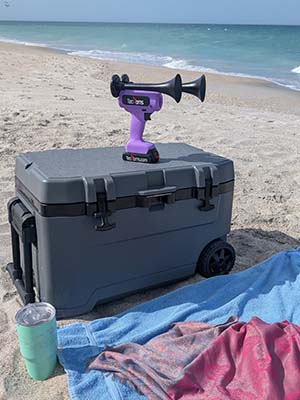 Purple TecHorn 2-Horn – handheld train horn with 20 V lithium battery, sitting on a beach cooler in the sand near the shoreline, showcasing its bold color and portable design for beach days and outdoor fun