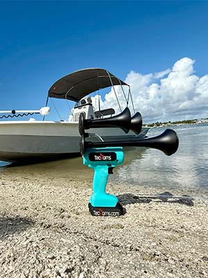 Teal TecHorn 2-Horn handheld train horn with 20 V lithium battery displayed on the sandy shore beside a boat, highlighting its coastal style and durable outdoor design