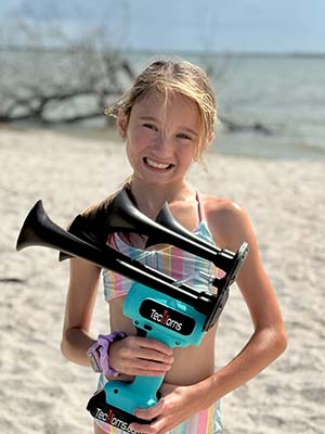Young girl holding a teal TecHorn 4-Horn handheld train horn with 20 V lithium battery on a sunny beach, showcasing fun, family, and Florida-built performance